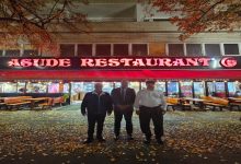 Görselde akşam saatlerinde bir restoranın önünde duran üç erkek görülüyor. Restoranın tabelasında kırmızı renkle “ASUDE RESTAURANT” yazıyor. Etrafta sonbahar yaprakları yerde birikmiş ve dış mekanda ahşap masalar bulunuyor. Arka planda restoranın içi ışıklarla aydınlatılmış şekilde görünüyor.