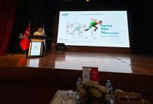 Sahne loş ışıklarla aydınlanmış, ağır ve saygın bir ortam var. Büyük bir projeksiyon perdesinde “Başarıya Giden Yol” başlıklı bir sunum görünüyor. Sol tarafta kürsünün arkasında bir kişi konuşma yapıyor; yanında Türk bayrağı ve İstanbul Ticaret Üniversitesi’nin flaması duruyor. Sahnenin önünde su şişeleri, çiçek aranjmanı ve küçük bayrakların bulunduğu bir masa dikkat çekiyor. Genel atmosfer, profesyonel ve akademik bir eğitim etkinliğini yansıtıyor.