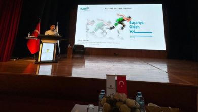 Sahne loş ışıklarla aydınlanmış, ağır ve saygın bir ortam var. Büyük bir projeksiyon perdesinde “Başarıya Giden Yol” başlıklı bir sunum görünüyor. Sol tarafta kürsünün arkasında bir kişi konuşma yapıyor; yanında Türk bayrağı ve İstanbul Ticaret Üniversitesi’nin flaması duruyor. Sahnenin önünde su şişeleri, çiçek aranjmanı ve küçük bayrakların bulunduğu bir masa dikkat çekiyor. Genel atmosfer, profesyonel ve akademik bir eğitim etkinliğini yansıtıyor.