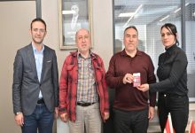 Görselde dört kişi yan yana ayakta poz veriyor. Sağdaki kadın, yanında duran erkeğe kırmızı bir kart ya da cihaz uzatıyor; erkek bunu elinde tutuyor. Ortam bir ofis gibi görünüyor; arkada cam bölmeler ve duvarda bir portre bulunuyor. Sol tarafta da iki erkek daha yer alıyor. Genel olarak resmi bir ziyaret veya teslim töreni havası var.