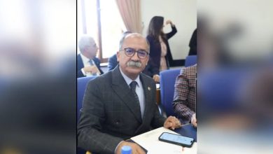 Görselde takım elbiseli, gözlüklü, bıyıklı bir erkek bir toplantı salonunda masada oturuyor. Önünde notlar ve telefonu var. Arkada başka katılımcılar da görünüyor.