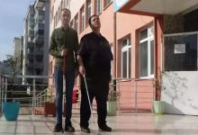 Fotoğrafta, bir okul binasının girişinde iki görme engelli öğretmen yan yana duruyor. Her ikisi de beyaz bastonlarıyla ayakta ve kendinden emin bir duruş sergiliyor. Soldaki öğretmen ince yapılı, yeşil bir gömlek giymiş; sağdaki öğretmen ise daha iri yapılı ve koyu renk bir kıyafet tercih etmiş. Arka planda okulun pembe tonlarındaki dış cephesi, pencereler ve giriş kapısı görünüyor. Güneşli bir hava, zemine düşen ışık ve gölgelerle belli oluyor. Sahne genel olarak güçlü, ilham veren bir birliktelik hissi taşıyor.