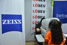 Görselde bir göz tarama etkinliği görülüyor. Sağ tarafta turuncu tişört giymiş bir çocuk, bir göz ölçüm cihazına bakıyor. Cihazın karşısında oturan görevli, ölçümü gerçekleştiriyor. Arka planda LÖSEV’in dikey afişi, yanında ZEISS ve Beta Optik logoları yer alıyor. Etkinlik ortamı oldukça düzenli; sağlık taramasına odaklanan, çocukların görme sağlığını destekleyen bir çalışma yürütüldüğü hissi veriyor.