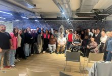 Görselde, geniş bir grup bir araya gelmiş şekilde poz veriyor. Farklı yaş ve özelliklerde birçok kişi ayakta veya sandalyelerde oturarak gülümsüyor. Ortam modern bir iç mekân; tavan aydınlatmaları, havalandırma boruları ve ahşap zemin dikkat çekiyor. Genel atmosfer sıcak, kapsayıcı ve topluluk ruhunu yansıtan bir buluşma izlenimi veriyor.