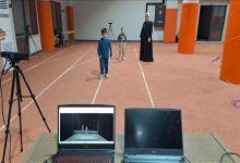 Geniş bir iç mekânda atletizm pistlerini andıran çizgiler var. Ortada, farklı yaşlarda iki çocuk ve arkalarında siyah uzun elbiseli bir yetişkin kadın ayakta duruyor. Ön planda bir masa üzerinde iki dizüstü bilgisayar bulunuyor. Bilgisayarlardan birinin ekranında, hareket yakalama/simülasyon benzeri bir görüntüyü gösteren bir sahne açık.