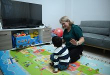 Bir oturma odasında, renkli oyun matı üzerinde diz çöken bir yetişkin, karşısında oturan küçük bir çocukla oyun oynuyor; etrafta oyuncaklar, arkada televizyon ve koltuk var.