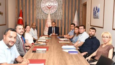 Bir toplantı salonunda uzun bir masa etrafında oturan dokuz kişi kameraya bakarak poz veriyor. Masanın başında bir yönetici yer alırken, arka duvarda Bakanlık logosu ve Türk bayrağı bulunuyor. Görsel, resmi bir değerlendirme veya koordinasyon toplantısını yansıtıyor.