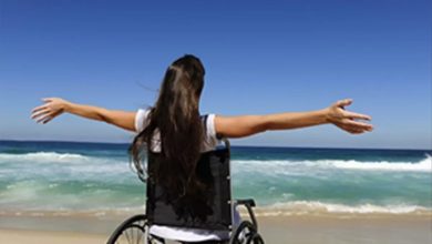 Bu fotoğrafta, tekerlekli sandalyede oturan uzun saçlı bir kadın deniz kenarında kollarını iki yana açmış şekilde poz veriyor. Kadının yüzü görünmüyor; kamera arkadan çekim yapmış. Önünde mavi gökyüzü, dalgalar ve sahil uzanıyor