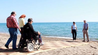 Bu fotoğrafta, kumsalda yerleştirilmiş ahşap bir yürüyüş yolunda yürüyen ve tekerlekli sandalye kullanan bireyler görülüyor. Yol, kumun üstüne serilerek hem engelli bireylerin hem de yaşlı ya da hareket kabiliyeti sınırlı kişilerin denize ulaşımını kolaylaştıracak şekilde tasarlanmış. Ön planda bir kişi elektrikli tekerlekli sandalyesiyle ilerliyor; yanında ona eşlik eden kişiler var. Arka planda ise deniz kenarında duran iki kişi görülüyor. Hava açık ve güneşli, deniz ise sakin.