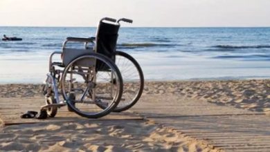 Bu fotoğrafta, bir plajda kumsalın üzerine yerleştirilmiş ahşap bir yürüyüş yolunun sonunda boş bir tekerlekli sandalye görülüyor. Sandalyenin hemen yanında bırakılmış terlikler dikkat çekiyor. Arka planda sakin bir deniz manzarası ve denizde yüzen insanlar yer almakta. Görsel, erişilebilirlik açısından olumlu bir örnek sunuyor; çünkü kumda tekerlekli sandalye ile ilerlemeyi kolaylaştıracak bir platform mevcut.