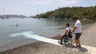 Bu fotoğraf, engelli bireylerin denize erişimini kolaylaştırmak amacıyla yapılmış özel bir yürüyüş yolunu gösteriyor. Sağ tarafta bir kişi, tekerlekli sandalyedeki bir bireyi kıyıya kadar getiriyor. Yol, tekerlekli sandalye ile rahat hareket edilebilecek şekilde denizin içine kadar uzanıyor. Arka planda tekneler, yüzen insanlar ve doğayla iç içe bir koy manzarası yer almakta. Fotoğraf, fiziksel engelleri aşmaya yönelik turizm altyapısının geliştiğini ve kapsayıcılığı temsil ediyor.