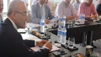 Bu fotoğraf bir toplantı ortamını göstermektedir. Masalarda su şişeleri, not defterleri, cep telefonları, kalemler ve tabaklarda sunulmuş yiyecekler yer almaktadır. Ön planda takım elbiseli bir adam not alırken görülüyor. Arka planda ise başka kişiler de masada oturmakta ve toplantıya katılmaktadır. Ortam resmi ve profesyonel bir izlenim vermektedir.