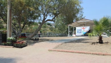 Bu fotoğraf, açık alanda çekilmiş ve “Özgür Plaj” tabelasının yer aldığı bir sahneyi göstermektedir. Sağ tarafta, üstü kapalı bir alanın girişinde büyük bir “Özgür Plaj” pankartı asılmış. Sol ve arka kısımlarda ağaçlar ve peyzaj düzenlemeleri görülüyor. Zeminde taş döşeme yollar, yanlarda çiçek kasaları ve yeni ekilmiş alanlar yer almakta. Görünen alan, engelli bireyler için erişilebilir şekilde düzenlenmiş bir plaj alanının girişini temsil ediyor.