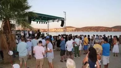 Fotoğrafta deniz kenarında düzenlenen bir etkinlik görülüyor. Ön planda ve orta alanda birçok insan sahilde toplanmış. Katılımcıların bazıları ayakta sohbet ediyor, bazıları ise sahnedeki etkinliği izliyor. Sol tarafta bir palmiye ağacı yer alıyor. Deniz kıyısında kurulan yeşil tente ile kaplı sahnede müzisyenler veya konuşmacılar bulunuyor. Sahnenin üstünde ışıklandırma ve ses sistemleri dikkat çekiyor. Arka planda deniz ve kıyıya paralel uzanan tepeler görülüyor. Hava açık ve gün batımı saatlerine yakın olduğu anlaşılıyor. Genel olarak yaz akşamında düzenlenen keyifli ve sosyal bir açık hava etkinliğini yansıtıyor.