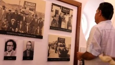 Fotoğrafta bir müze ortamı görülüyor. Sağ tarafta beyaz gömlek giymiş bir adam, duvara asılı bir pano üzerindeki siyah-beyaz fotoğraflara dikkatle bakıyor. Panoda, üzerinde "Sünger Kooperatifi" yazan eski dönem fotoğrafları bulunuyor. Fotoğraflar, geçmişteki süngercilik faaliyetlerini ve bu işle uğraşan insanları yansıtıyor. Ayrıca panoda iki adet portre fotoğrafı da yer almakta. Mekânın duvarı beyaz renkli ve duvarda yuvarlak bir pencere veya açıklık bulunuyor. Genel olarak tarihî ve kültürel bir atmosfer hakim.