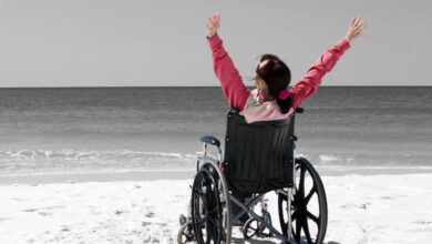 Kumsalda tekerlekli sandalye üzerinde oturan engelli birey yüzü sahile doğru dönük bir şekilde kollarını yukarı doğru ve her iki yana doğru açarak mutluluğunu dile getiriyor.