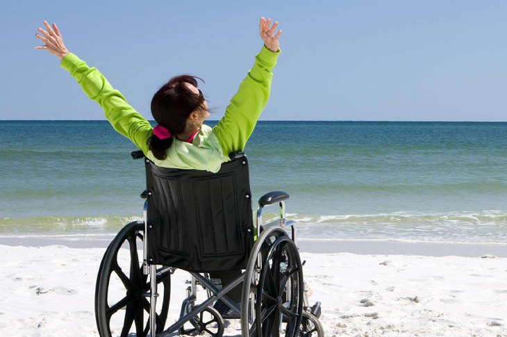 Tekerlekli sandalyede bulunan kadın sahilde iki kolunu havaya kaldırıyor.