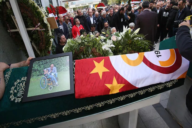 Yavuz Kocaömer'in cenazesine Galatasaray bayrağı asılı ve engelli bir çocukla çekilen fotoğrafı bulunuyor, cemaat törende bekliyor.