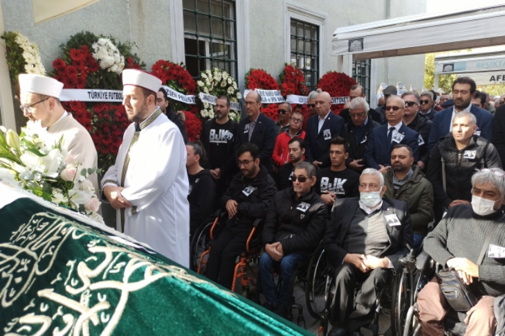 Cenaze namazı kılınıyor ve iki imamın arkasında tekerlekli sandalyeli kişiler ile cemaat törende bulunuyor.
