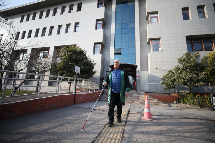 Görme Engelli Avukat Salih Mehmet Aslan, elinde beyaz baston tutarak sarı renkli hissedilebilir yolda yürüyor.