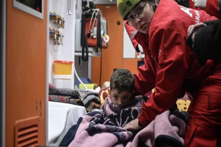 3 yaşındaki Muhammed Yusuf Tanık ambulans içinde battaniyeye sarılı durumda oturuyor ve yanında kadın arama kurtarma personeli bulunuyor.