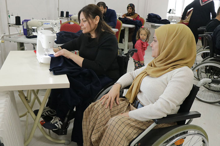 Tekerlekli sandalye kullanan bedensel engelli üye dikiş makinesi ile dikiş yaparken tekerlekli sandalyeli iş arkadaşı kendisini izliyor.