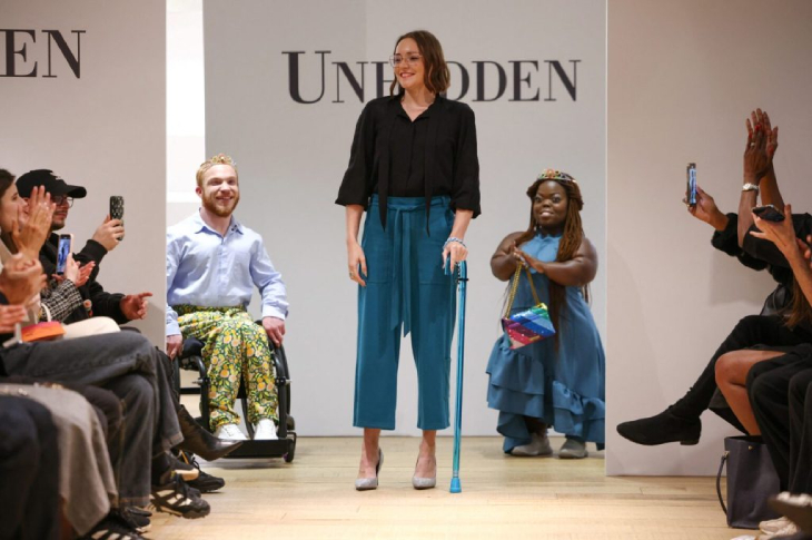 Podyumun ortasında mavi renkli el değneği tutan siyah kazaklı, açık petrol mavisi renkli model kadın bulunurken arkasında sol tekerlekli sandalyeli erkek model ile Afrika kökenli kısa boylu model kadın bulunuyor.