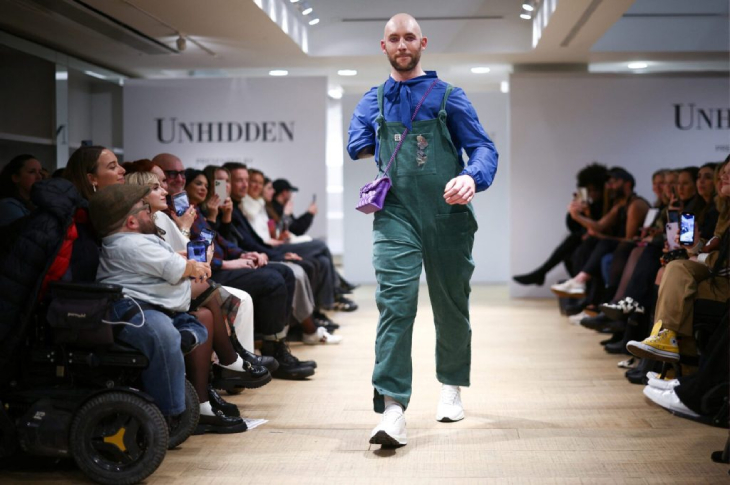 Sağ kolu ampute olan erkek model üstünde mavi kazak, bahçıvan pantolon ve mor çanta takılı olarak podyumda yürüyor.