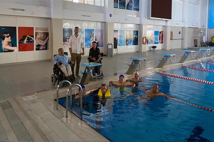 Havuz kenarındaki akülü sandalyeli ve havuz içindeki engelli bireyler objektife yansıyor.
