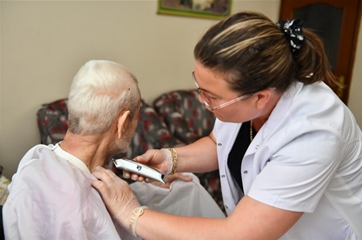 Yaşlı adamın sakal tıraşını yapan bir bayan objektife yansıyor.