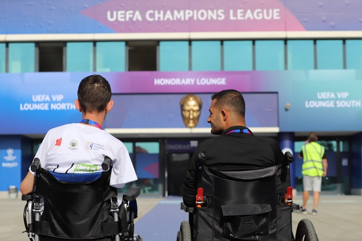 Stadyumun giriş kapısı önünde, akülü tekerlekli sandalyeli 2 birey objektife yansıyor.