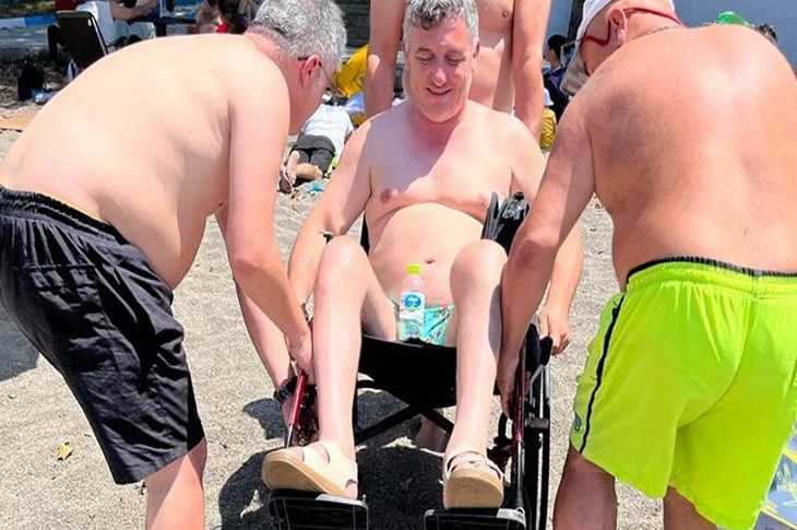 Kumsaldaki tekerlekli sandalyeli bireye 2 gönüllü bireyin yardımcı olduğu ana dair bir görüntü objektife yansıyor.