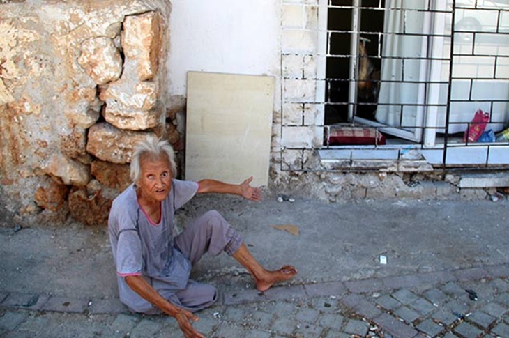 Evinin önünde, yerde oturan yaşlı kadının çaresiz durumu objektife yansıyor.