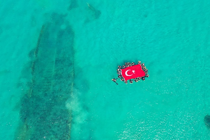 Dalış alanında, su yüzeyinde dev bayrak açan ekip objektife yansıyor.