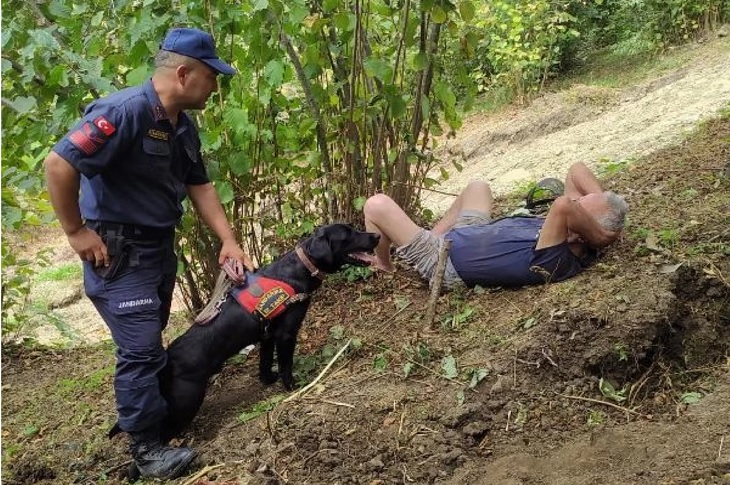 Jandarma görevlisi ve iz takip köpeği, yerde uzanan alzheimer hastası adamla birlikte objektife yansıyor.