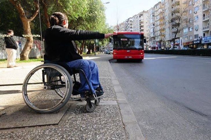 Yolda, özel halk otobüsüne durması için el uzatan bir tekerlekli sandalyeli birey objektife yansıyor.