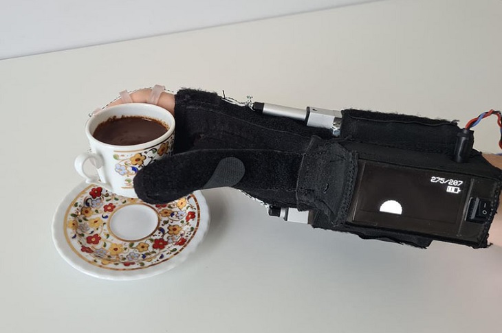 Dış iskelet ürününü giyerek içi dolu kahve fincanını tutan bir el resmi objektife yansıyor.