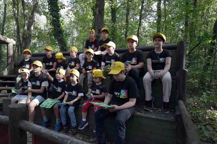 Ormanlık bölgede düzenlenen etkinlikte sıralı halde oturan çocuklar objektife yansıyor.