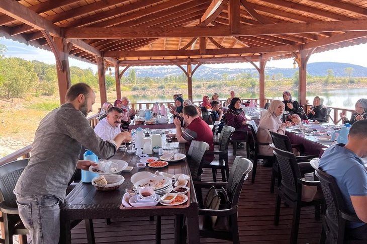 Doğa manzaralı mekanda, masalarında yemek yiyen çok sayıda katılımcı objektife yansıyor.