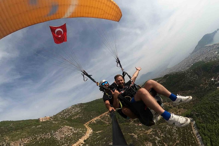 Bayrak asılı bir yamaç paraşütü ile havada süzülen 2 birey objektife yansıyor.