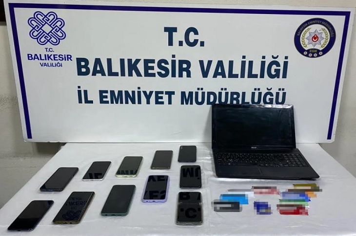İl Emniyet Müdürlüğü tarafından masada sergilenen cep telefonları, banka kartları ve dizüstü bilgisayar objektife yansıyor.