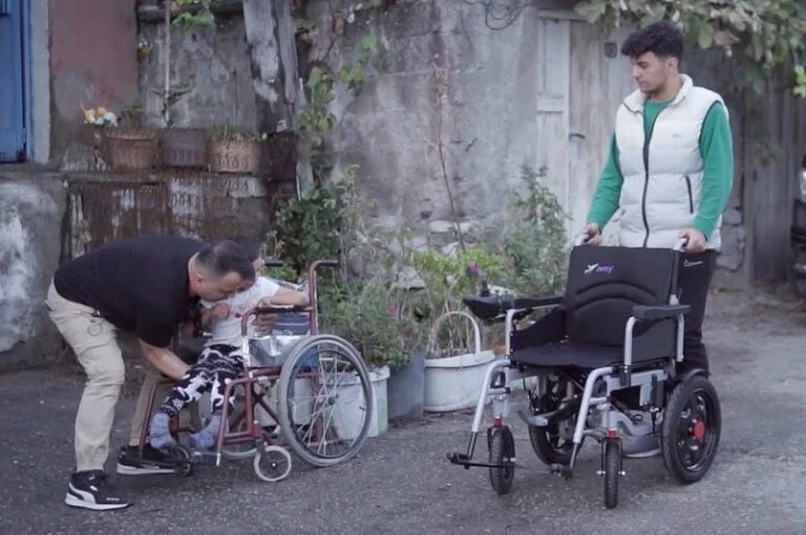 Mustafa'yı tekerlekli sandalyesinden yeni akülü sandalyesine taşıyan 2 kişi objektife yansıyor.