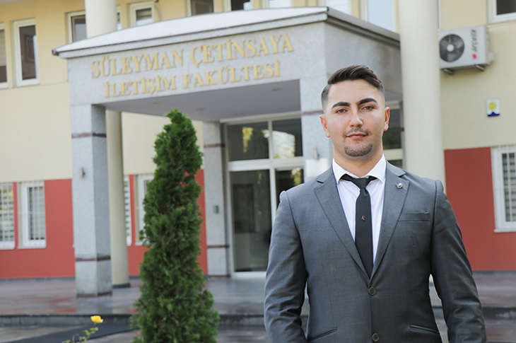 Yahya Mert, resmi giyimiyle iletişim fakültesi ana giriş kapısı önünde objektife yansıyor.