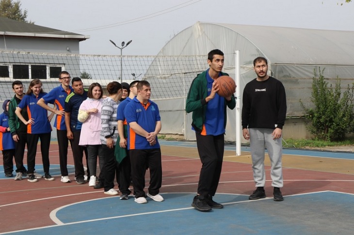 Tek sıra halinde, basketbol potasına sırayla atış yapan engelli bireyler objektife yansıyor.