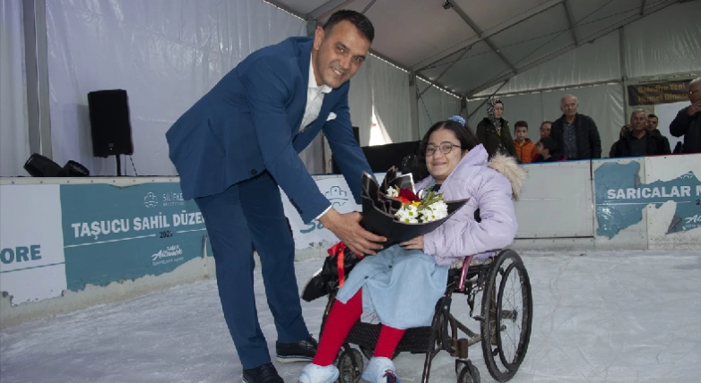 Tekerlekli sandalyeli engelli kız çocuğuna belediye yetkilisi buz pateni pistinde çiçek uzatırken