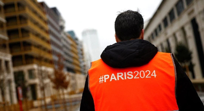 Paris2024 yazılı yeleğiyle görevli erkek sırtı dönük paris sokaklarına bakarken