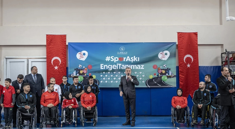 Afiş önünde bakan, sağ ve solunda engelli sporcular ile ayakta duruyor.