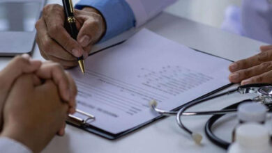 Beyaz bir önlük giymiş olan doktorun reçete yazarken, hastayla elleri masada durmaktadır.