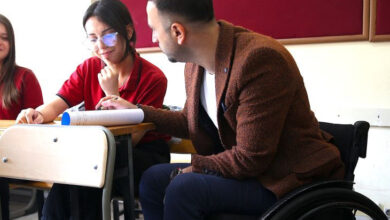 Tekerlekli sandalye kullanıcısı Ökkeş Aygün Öztürk, sırada oturan öğrencisinin yanında ders anlatıyor.