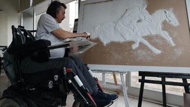 Tekerlekli sandalye üzerinde oturarak, elindeki resim boyaları ve tuval üzerine resim yapıyor.